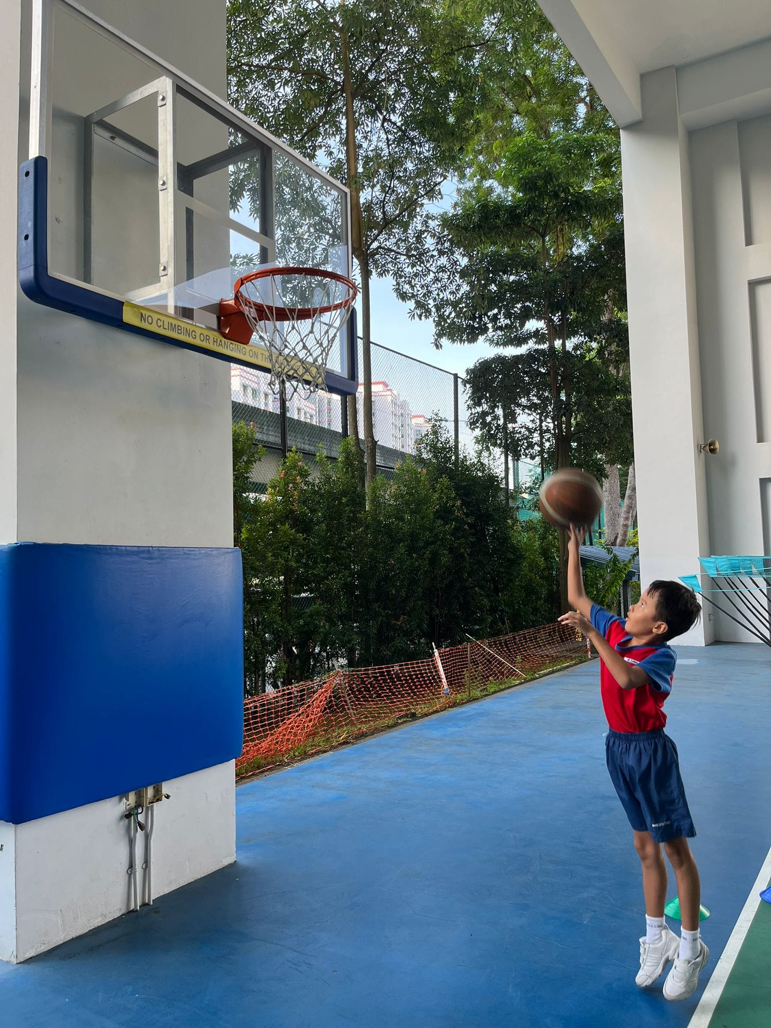 Young boy in red shirt shooting a basketball toward a wall-mounted hoop on a blue outdoor court.